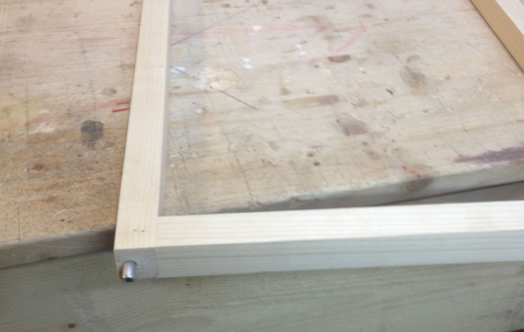 Close-up of a light wooden frame on a workbench. A metal pin is visible in one corner.