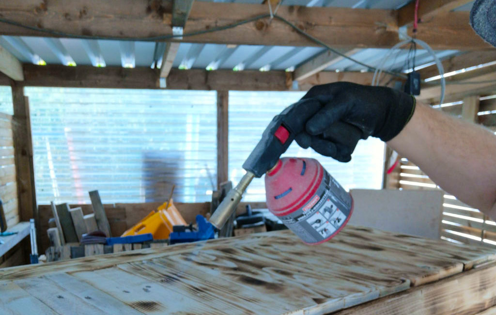 A person holds a blowtorch with a red gas cartridge and burns wood in a workshop.