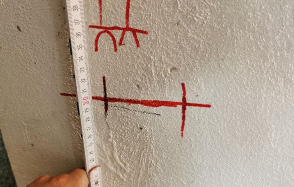 Eine Hand hält ein Messwerkzeug an eine Wand mit roten Markierungen für Bohrpunkte und Abstände.