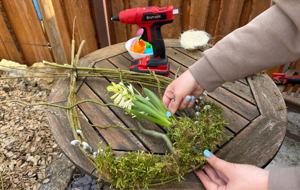 Hands placing flowers into a moss-covered wreath made of branches, with an Einhell hot glue gun behind.