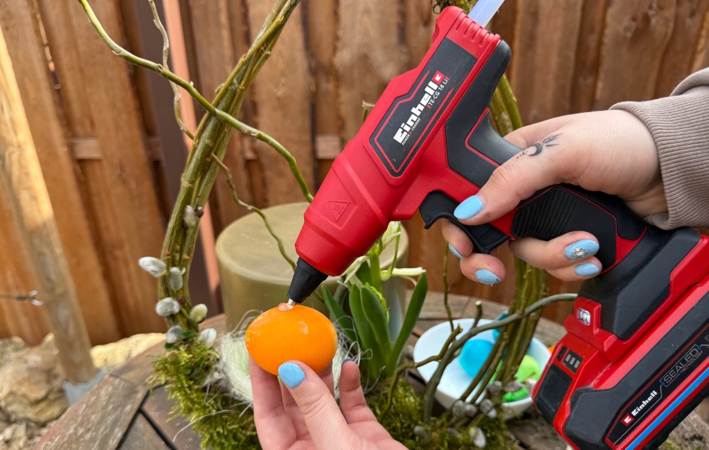 A colored egg is glued onto a decorative wreath using an Einhell hot glue gun.