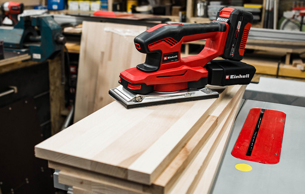 Einhell cordless sander placed on light wooden board in a workshop with tools in the background.