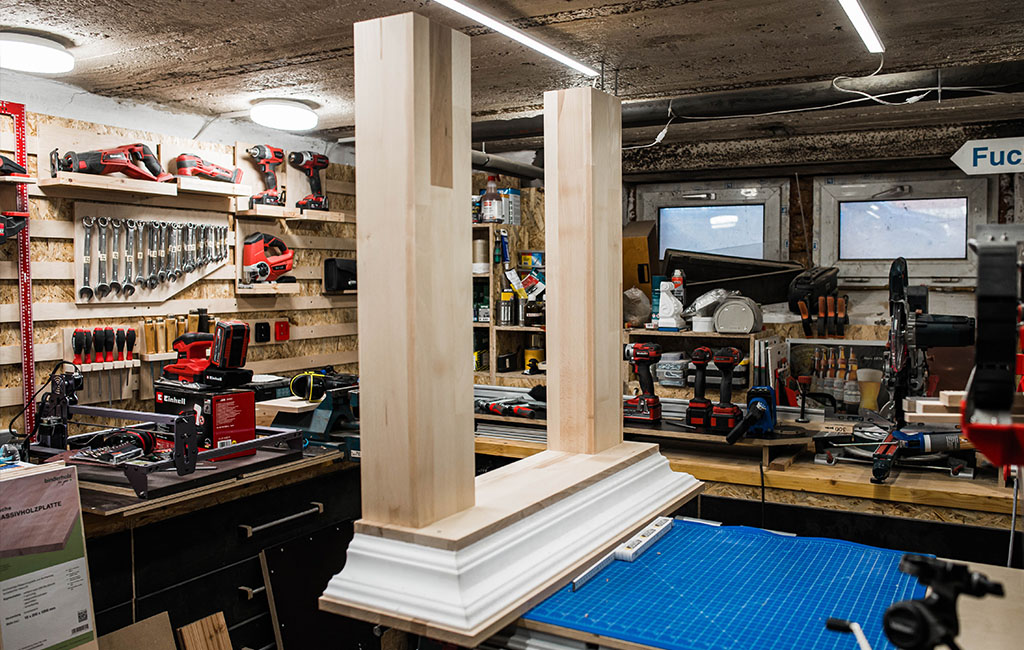 Two vertical wooden supports on a white base in a workshop with tool wall and machines.