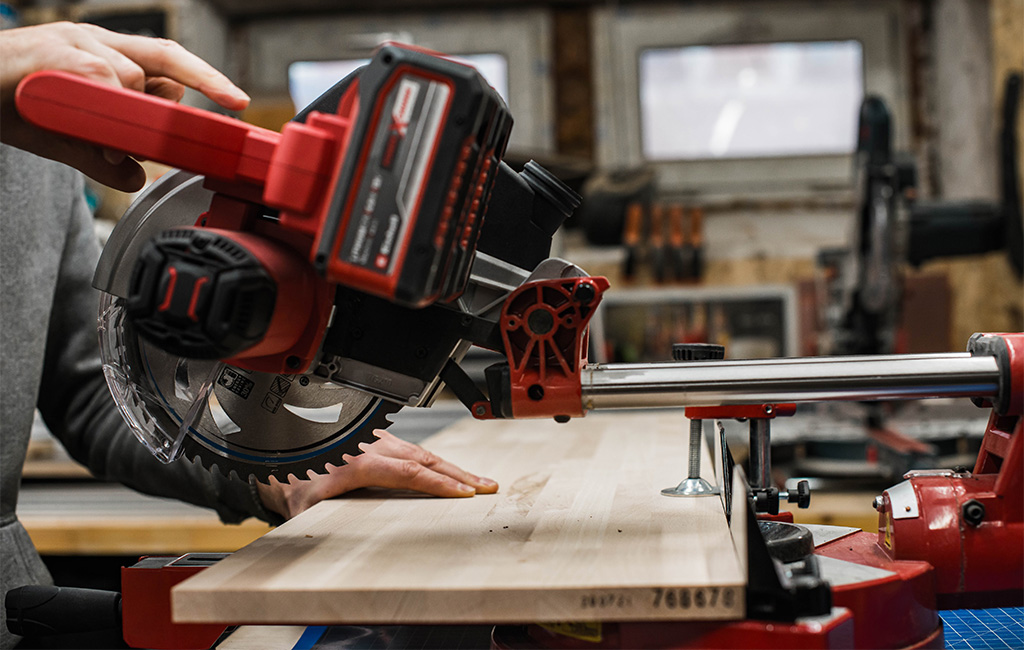 A person uses an Einhell cordless mitre saw to cut a light wooden board in the workshop.