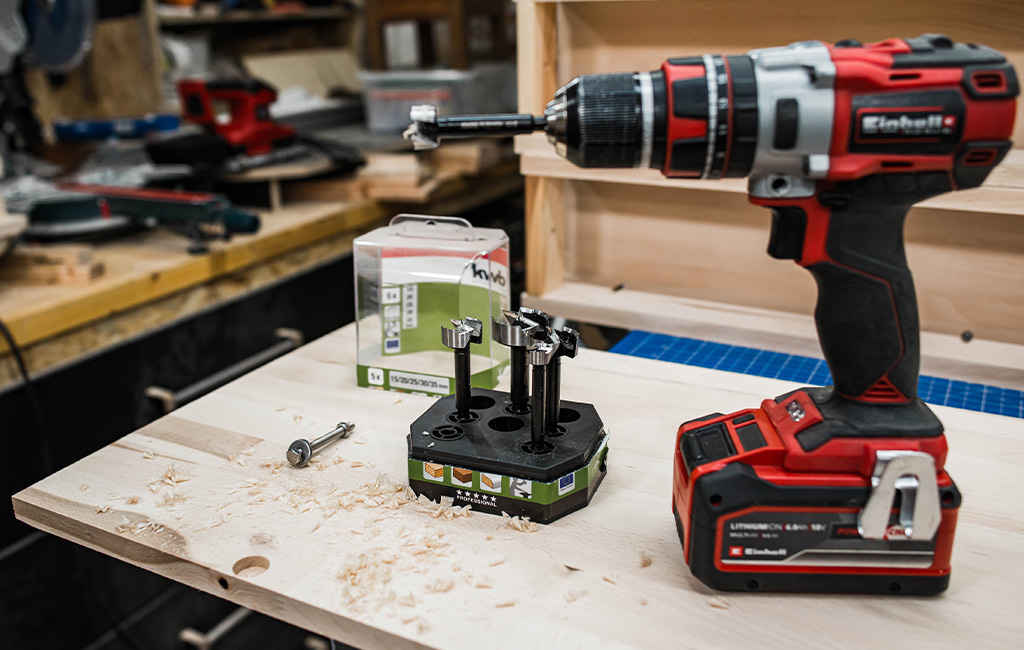 An Einhell cordless drill with Forstner bits and kwb packaging on a wooden board in the workshop.