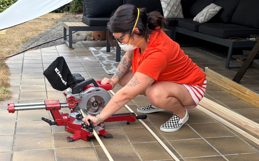 A woman cuts a narrow wooden strip with a red Einhell mitre saw on a patio.