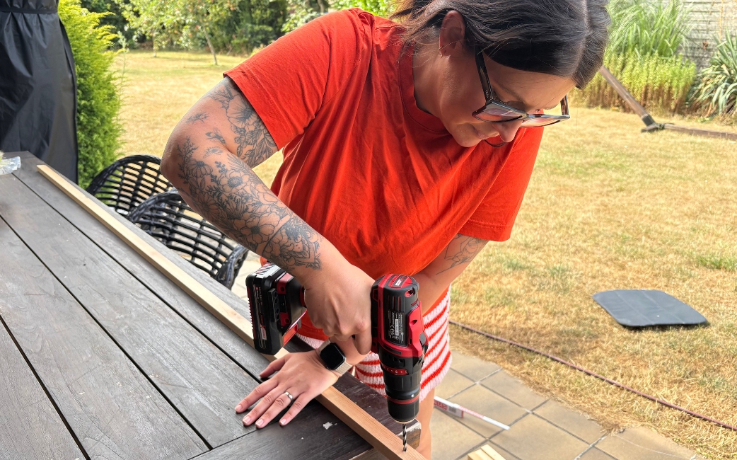 A woman drills a hole into a wooden strip with a red Einhell cordless drill driver.