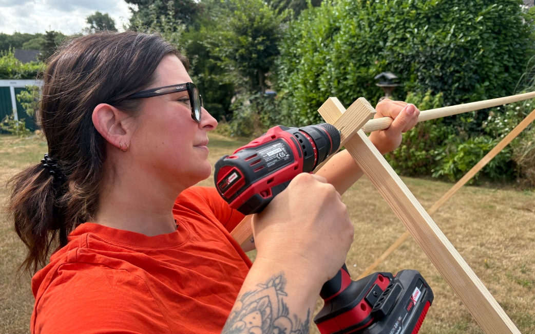 A woman uses a red Einhell cordless drill driver to attach a round wooden rod to an angled wooden frame.