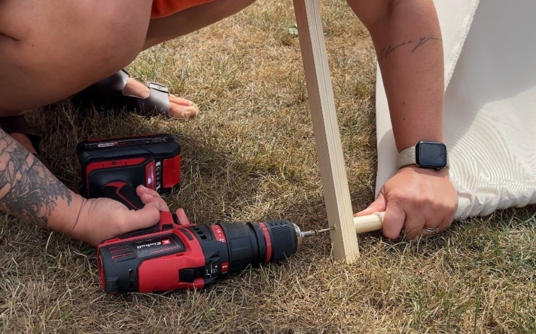 A woman uses a red Einhell cordless drill driver to attach a round wooden rod to the lower end of a wooden frame.