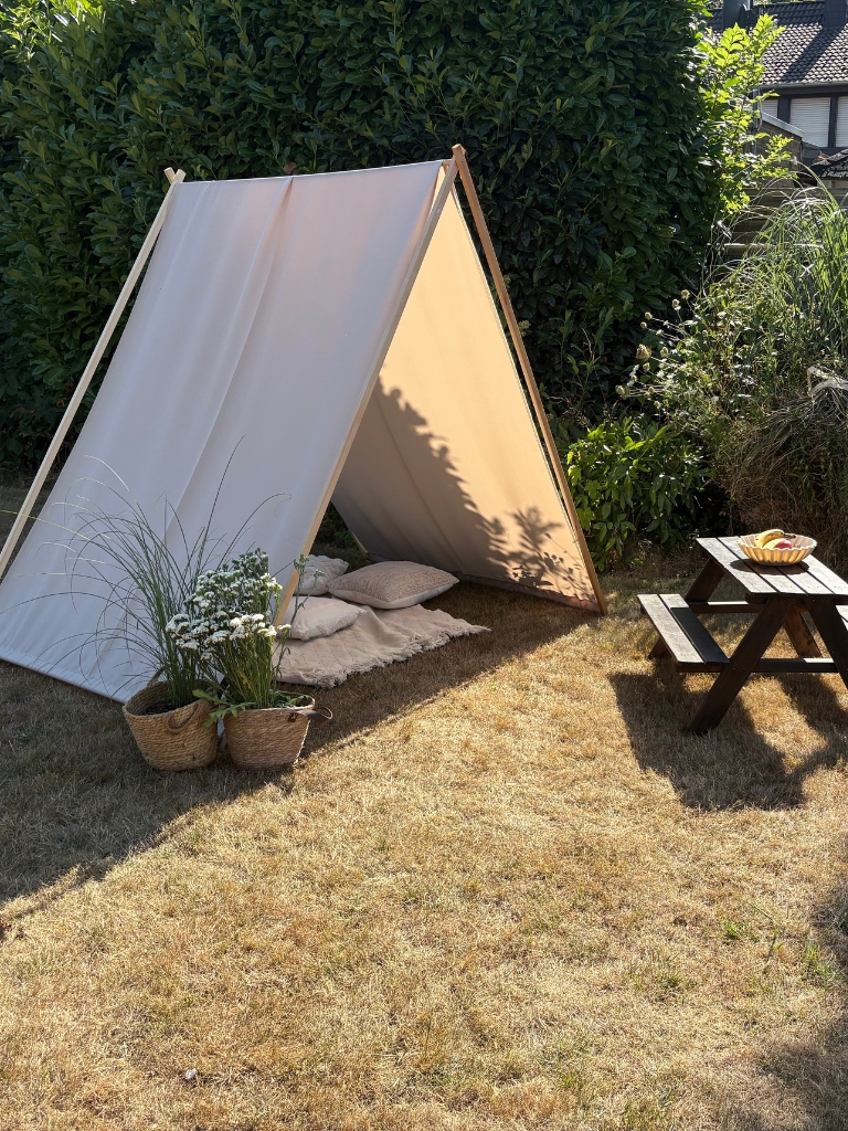 Beige fabric stretched over an A-shaped wooden frame as a sun shelter in a garden, with a picnic table and plants nearby.