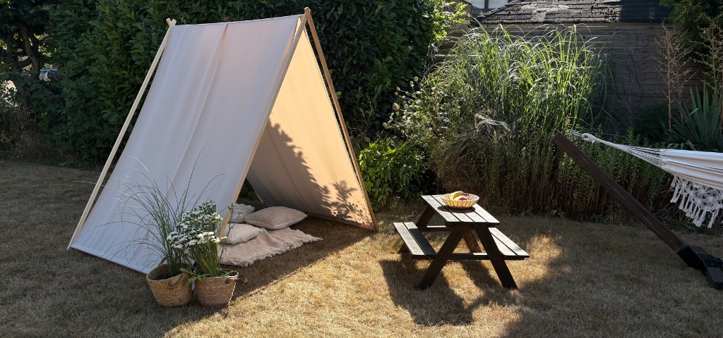 Beige fabric stretched over an A-shaped wooden frame as a sun shelter in a garden, with a picnic table and plants nearby.
