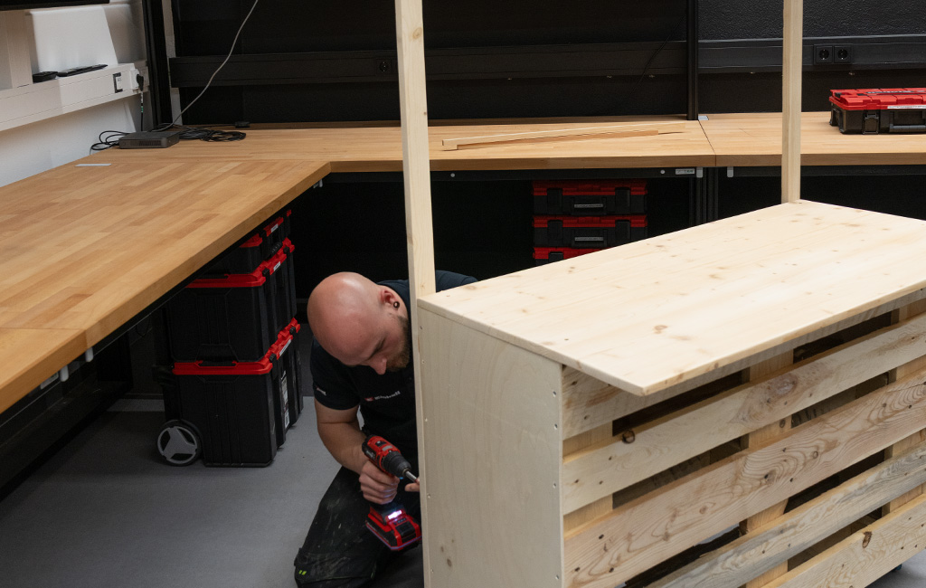 In einer Werkstatt montiert ein Mann mit einem Einhell Akku-Bohrschrauber die obere Holzkonstruktion an der fast fertigen DIY-Palettenbar.