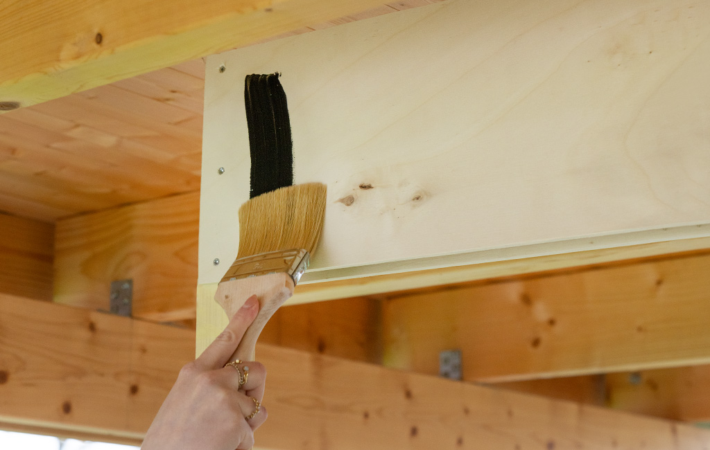 Eine Hand streicht mit einem breiten Pinsel schwarze Tafelfarbe auf ein helles Holzbrett der Bar-Konstruktion.