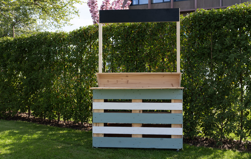 Die fertig aufgebaute, unbestückte Palettenbar mit grün-weiß lackierter Front und einem schwarzen Schild steht auf einer Rasenfläche vor einer Hecke.