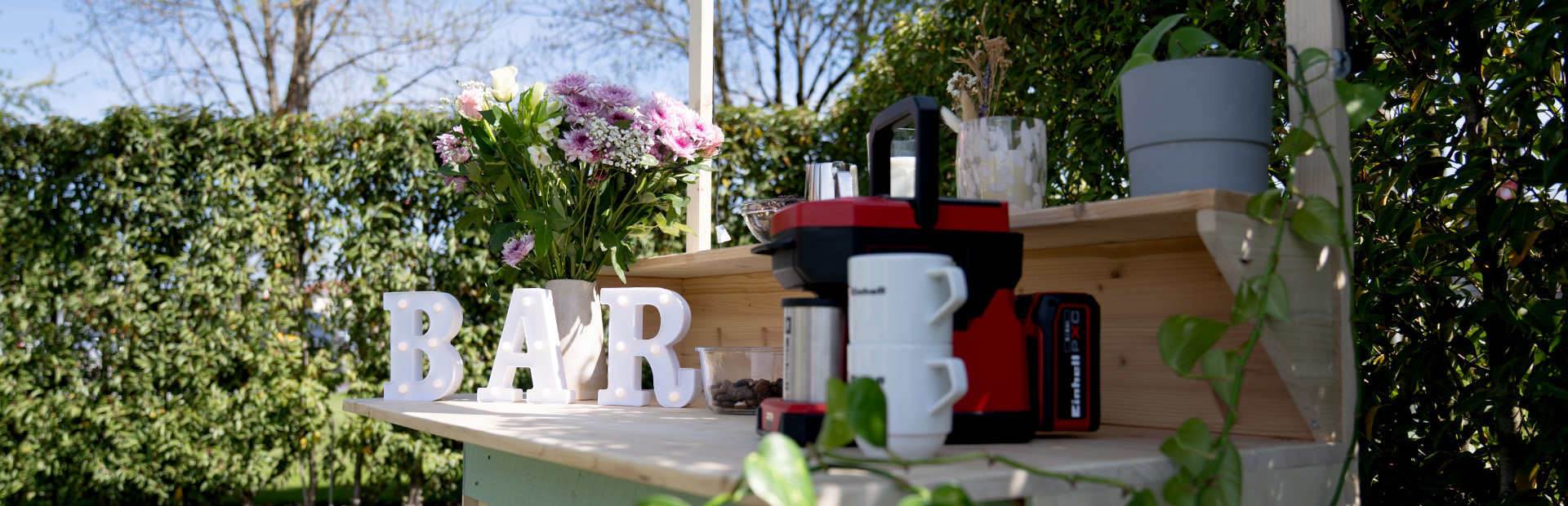 Eine fertig dekorierte DIY-Gartenbar aus Holzpaletten im Grünen mit beleuchteten Buchstaben, die das Wort „BAR“ formen, Blumen und einer roten Kaffeemaschine von Einhell.