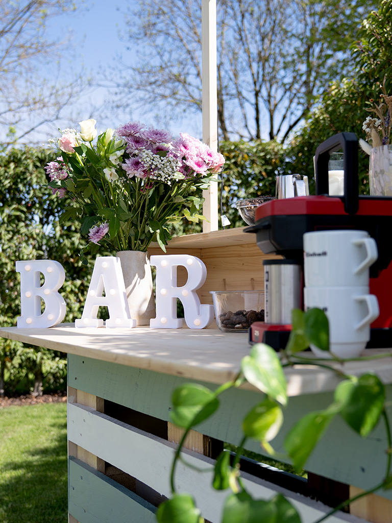 Eine fertig dekorierte DIY-Gartenbar aus Holzpaletten im Grünen mit beleuchteten Buchstaben, die das Wort „BAR“ formen, Blumen und einer roten Kaffeemaschine von Einhell.