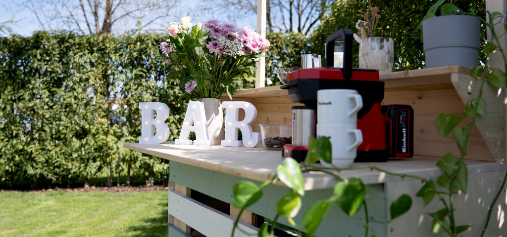 Eine fertig dekorierte DIY-Gartenbar aus Holzpaletten im Grünen mit beleuchteten Buchstaben, die das Wort „BAR“ formen, Blumen und einer roten Kaffeemaschine von Einhell.