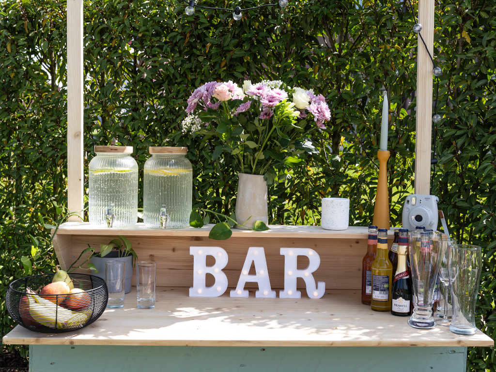 Eine fertig dekorierte DIY-Gartenbar aus Holzpaletten im Grünen mit beleuchteten Buchstaben, die das Wort „BAR“ formen, Blumen und einer roten Kaffeemaschine von Einhell.