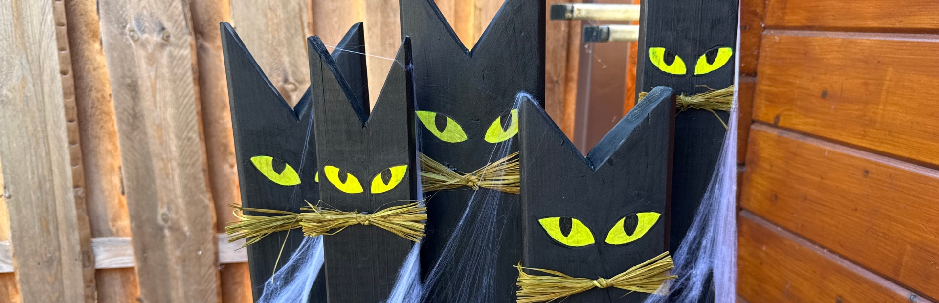 Fünf schwarze, katzenförmige Holzfiguren mit gelben Augen und Schnurrhaaren aus Stroh stehen auf einem gemeinsamen Holzsockel.