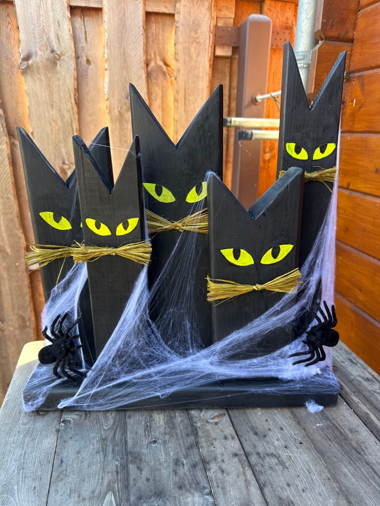 Fünf schwarze, katzenförmige Holzfiguren mit gelben Augen und Schnurrhaaren aus Stroh stehen auf einem gemeinsamen Holzsockel.