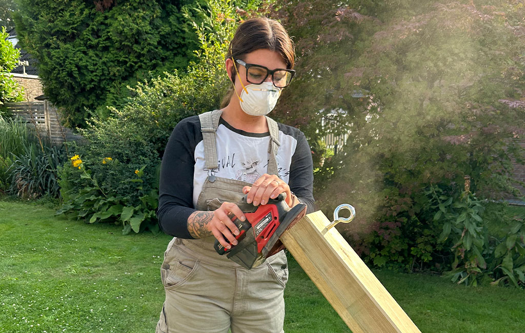 Eine Frau schleift einen Holzbalken mit einem Einhell Akku-Multischleifer; sie trägt Schutzbrille und Maske.