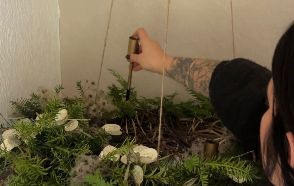 Eine Hand steckt eine braune Kerze in eine goldfarbene Halterung im Adventskranz.