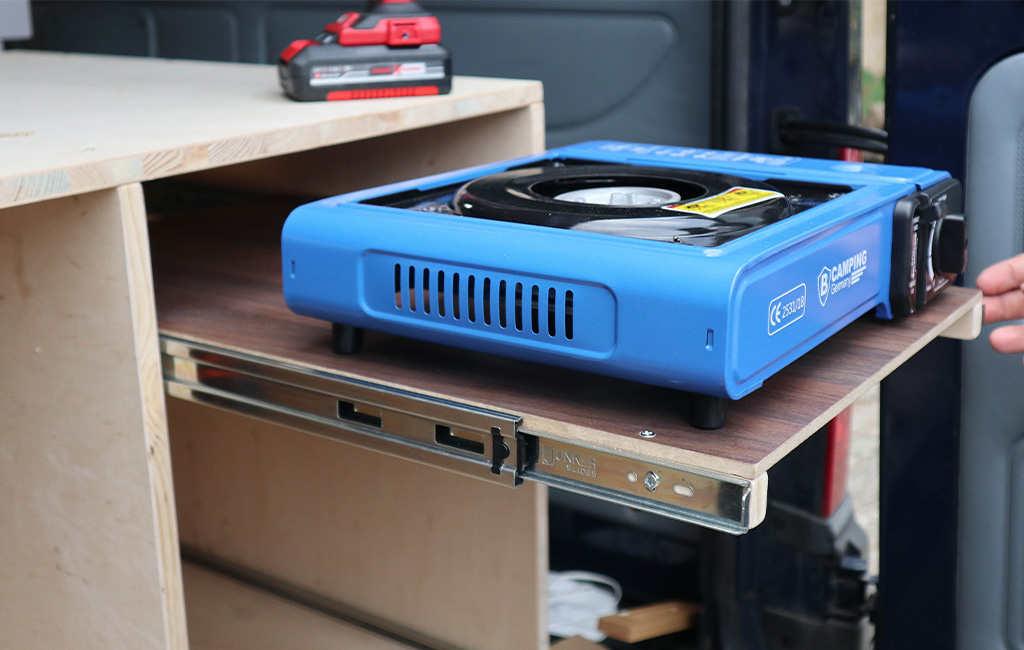 Blauer Campingkocher auf einem ausziehbaren Holzbrett mit Metallführung im Camper.