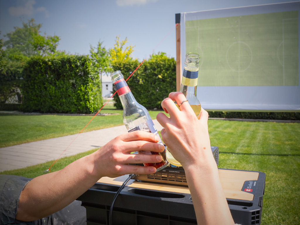 Zwei Hände stoßen im Vordergrund mit Getränkeflaschen an, während im Hintergrund ein Fußballspiel auf einer selbstgebauten Outdoor-Leinwand läuft.