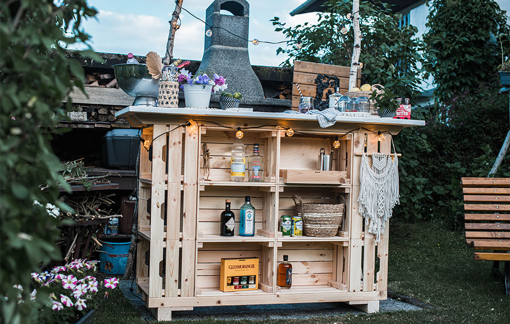 Die fertig gebaute Gartenbar steht dekoriert im Garten mit Getränken und Pflanzen.