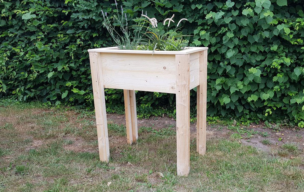 A tall, rectangular raised bed made of light wood with plants, standing on grass in front of a green hedge.