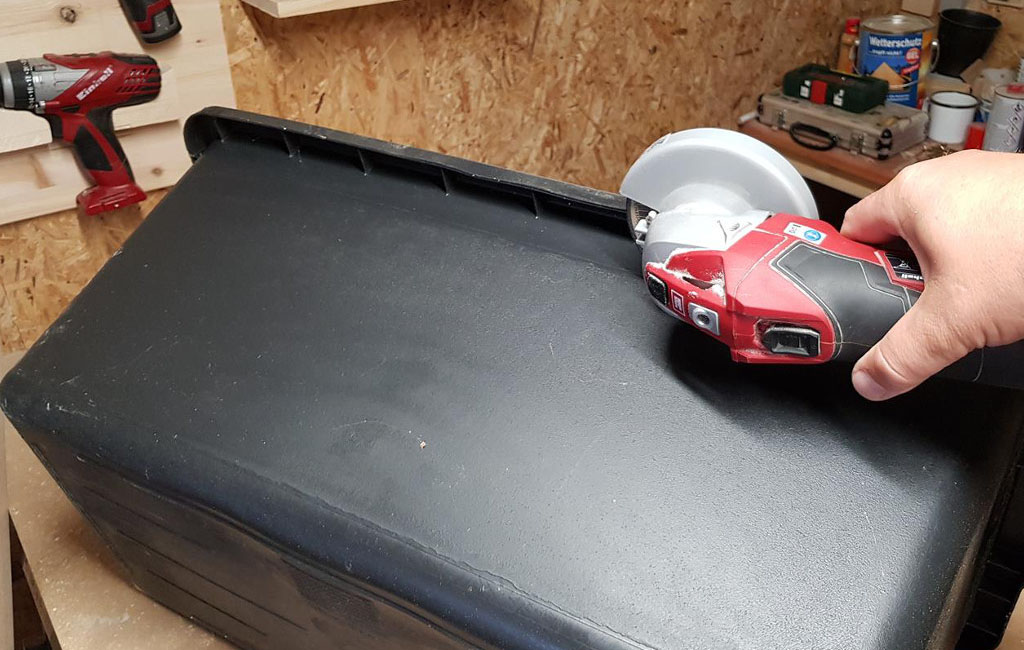 A hand holds an Einhell angle grinder and cuts the edge of a black plastic box.