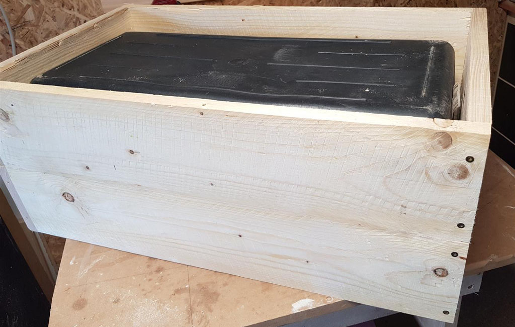 A black plastic box is placed inside a rectangular wooden frame made of light wood planks.