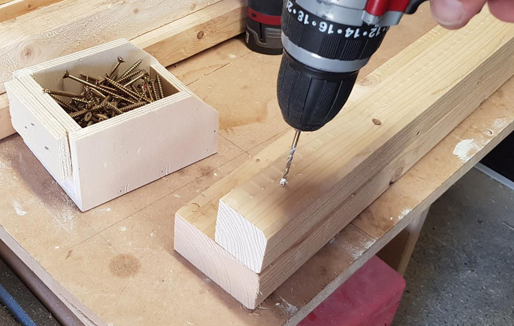 An Einhell cordless screwdriver drills a hole in a thick piece of wood, next to it is a box of screws.