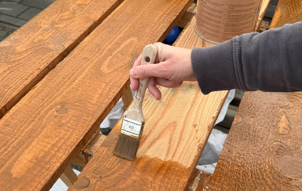 Eine Hand streicht mit einem Pinsel braune Lasur auf eine Holzplanke, daneben steht eine Farbdose.