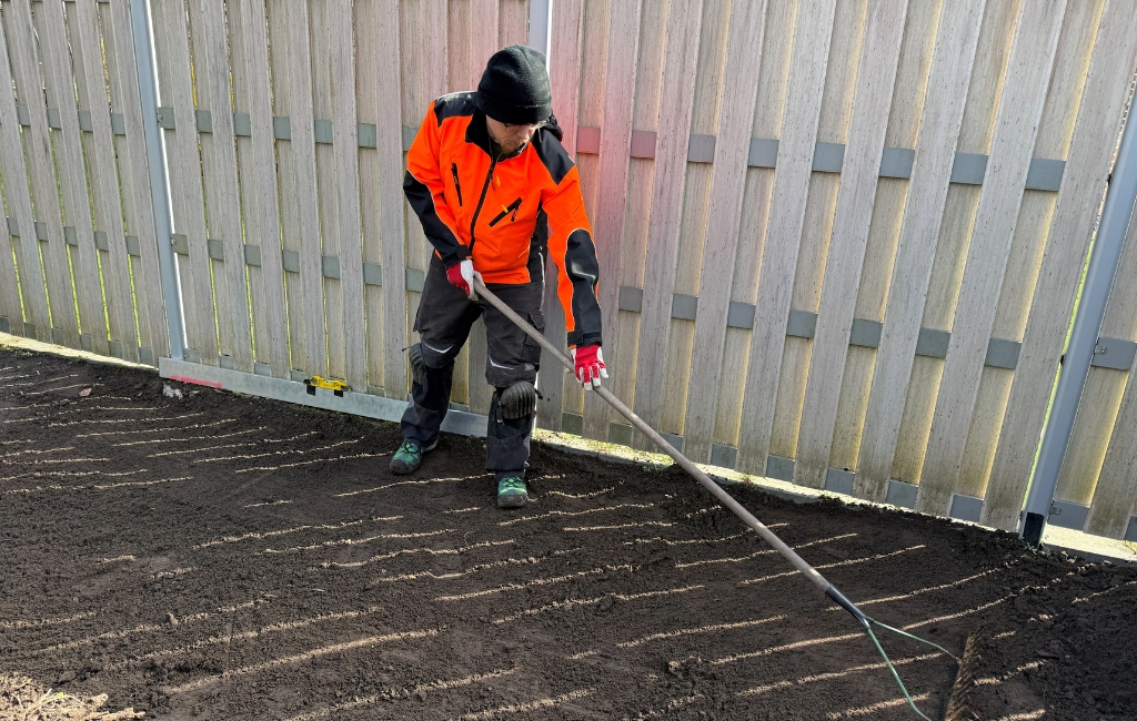 Ein Mann in Arbeitskleidung ebnet mit einem Rechen die dunkle Erde vor einem Holzzaun ein.