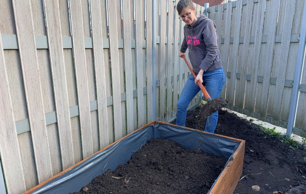 Eine Frau steht an einem Hochbeet und füllt mit einer Schaufel dunkle Erde in den mit Folie ausgekleideten Holzrahmen.