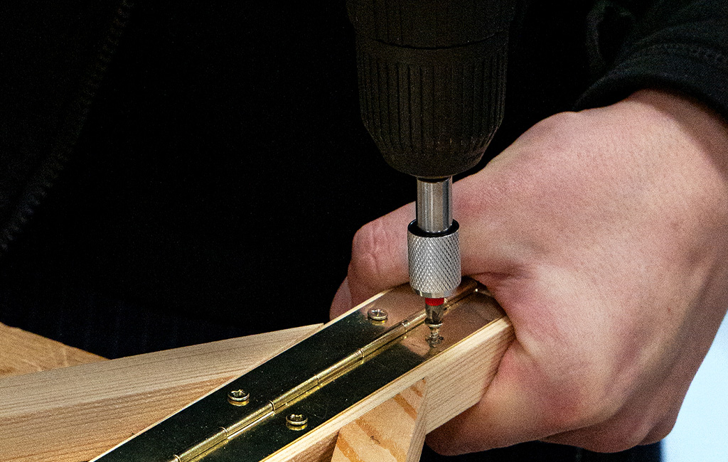 Eine Person schraubt mit einem Akkuschrauber ein Metallscharnier an zwei Holzleisten fest.