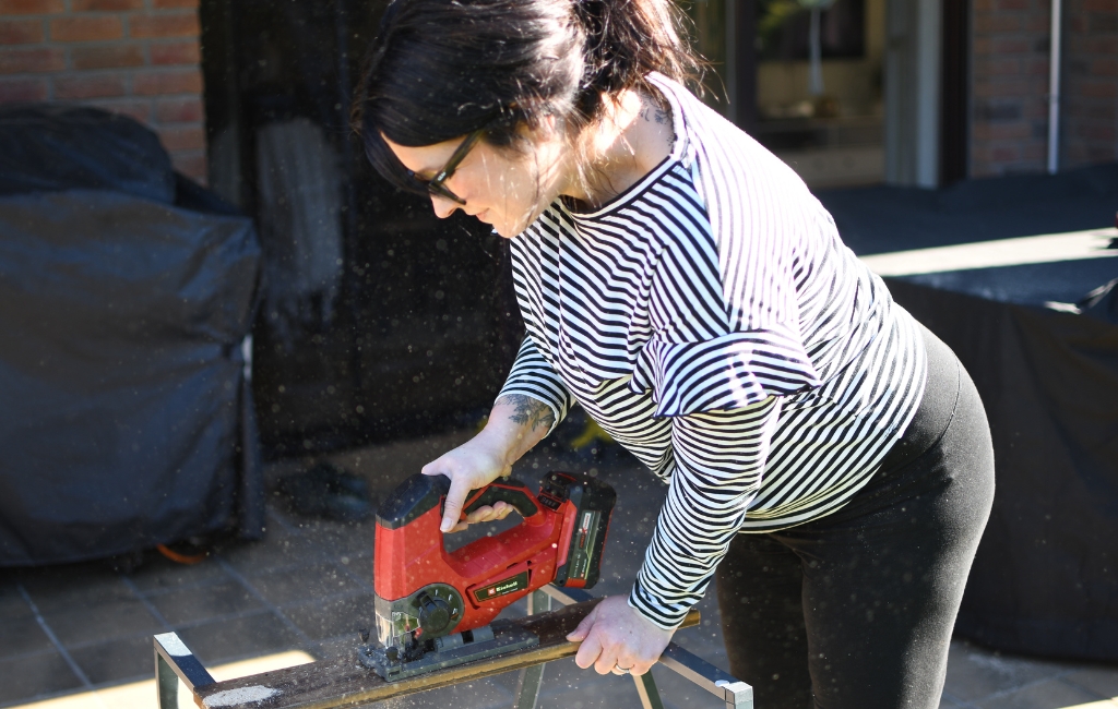 Person sägt Holz mit einer roten Akku-Stichsäge auf einem Arbeitstisch im Freien.