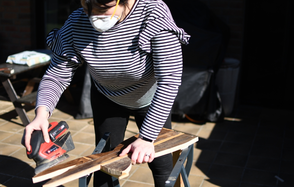 Person schleift ein Holzbrett mit Akku-Multischleifer auf einem Arbeitstisch im Freien.