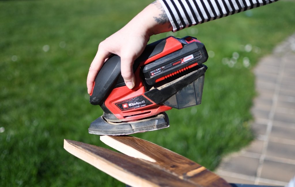 Hand schleift ein Holzbrett mit einem roten Akku-Multischleifer im Freien.