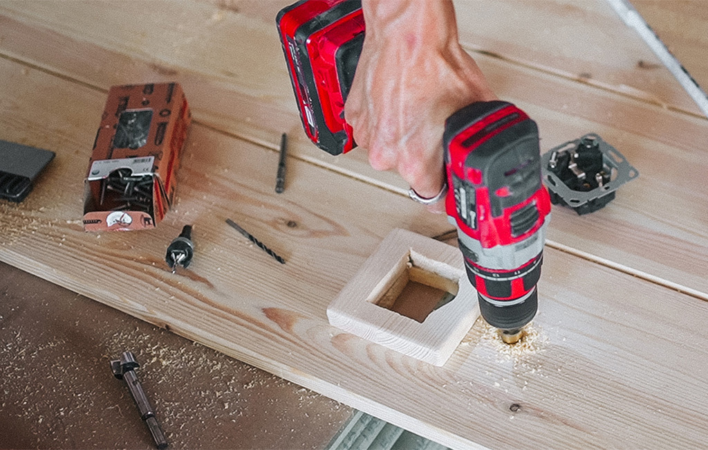 Eine Holzplatte wird mit einer Lochsäge für die Steckdose im Strandkorb bearbeitet.