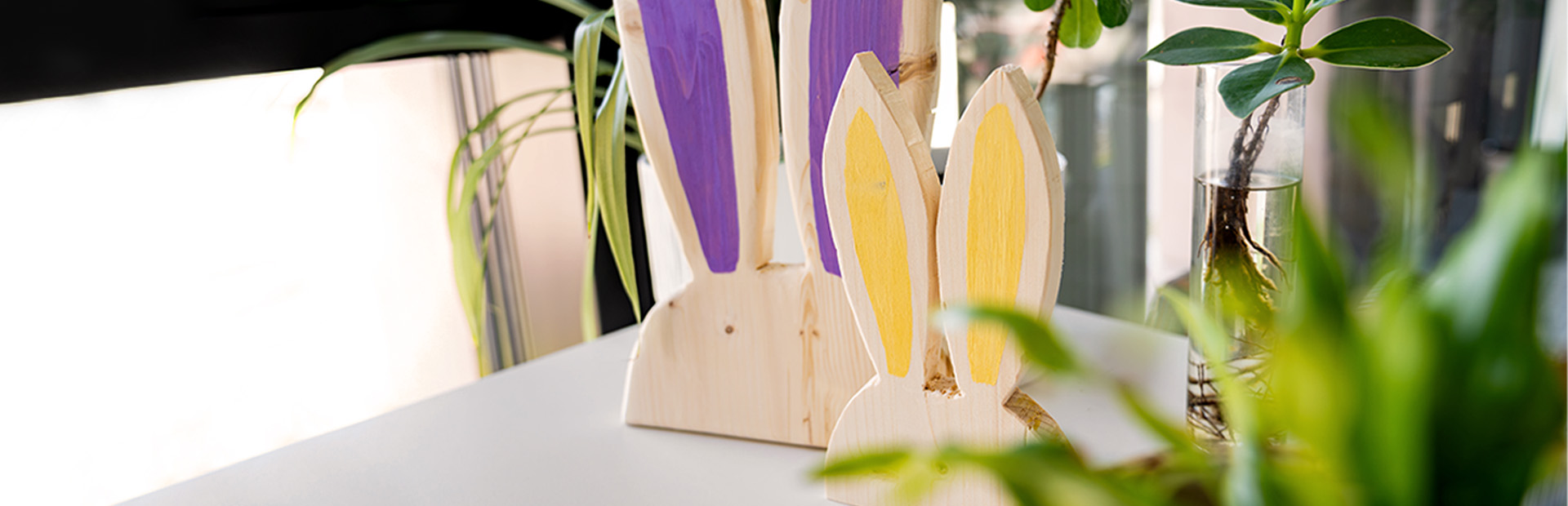 Zwei dekorative Holzhasen mit bemalten Ohren stehen auf einem Tisch neben Zimmerpflanzen.