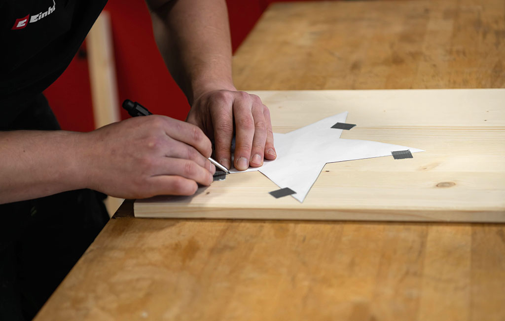 Hände zeichnen mit einem Stift einen Papierstern nach, der mit Klebeband auf einem Holzbrett fixiert ist.