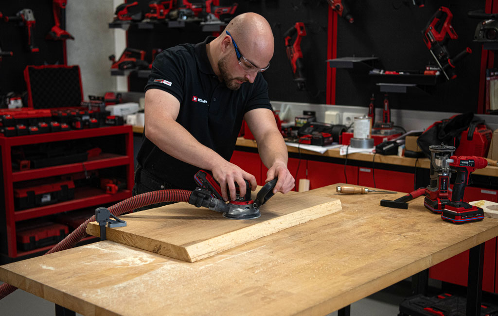 Eine Person schleift mit einem Exzenterschleifer eine große Holzplatte in einer Werkstatt.