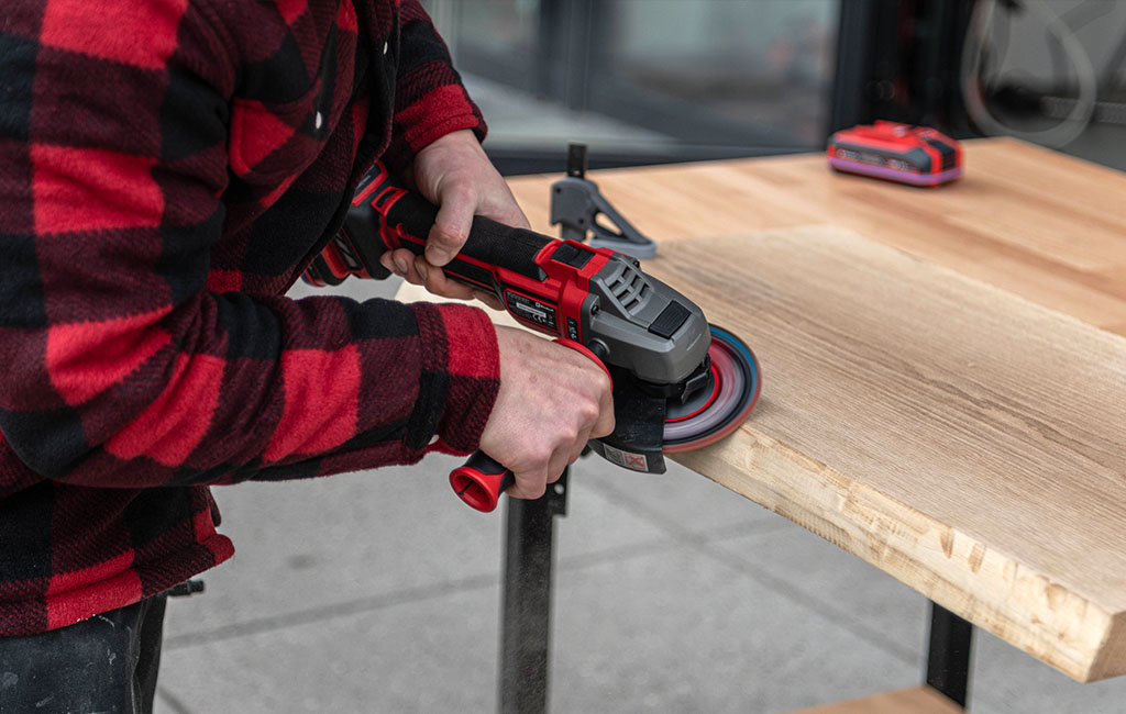 Eine Person schleift im Freien die Oberfläche einer Holzplatte mit einem Winkelschleifer.