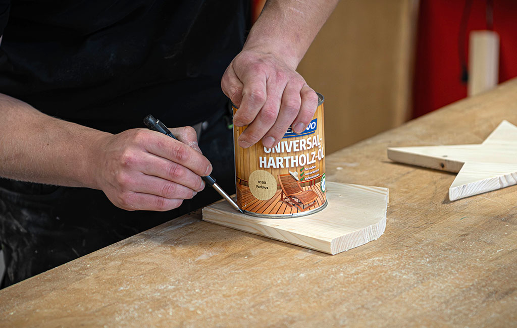 Eine Person nutzt eine Dose „Universal Hartholz-Öl“, um eine Markierung auf einem Holzstern zu setzen.