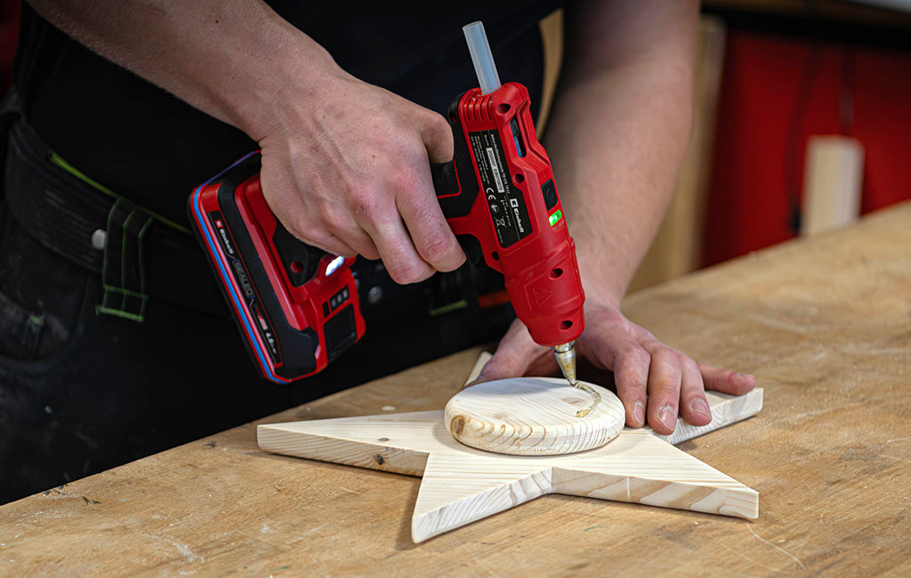 Eine Person trägt mit einer roten Einhell Heißklebepistole Klebstoff auf einen runden Holzteil auf, der auf einem Holzstern liegt.