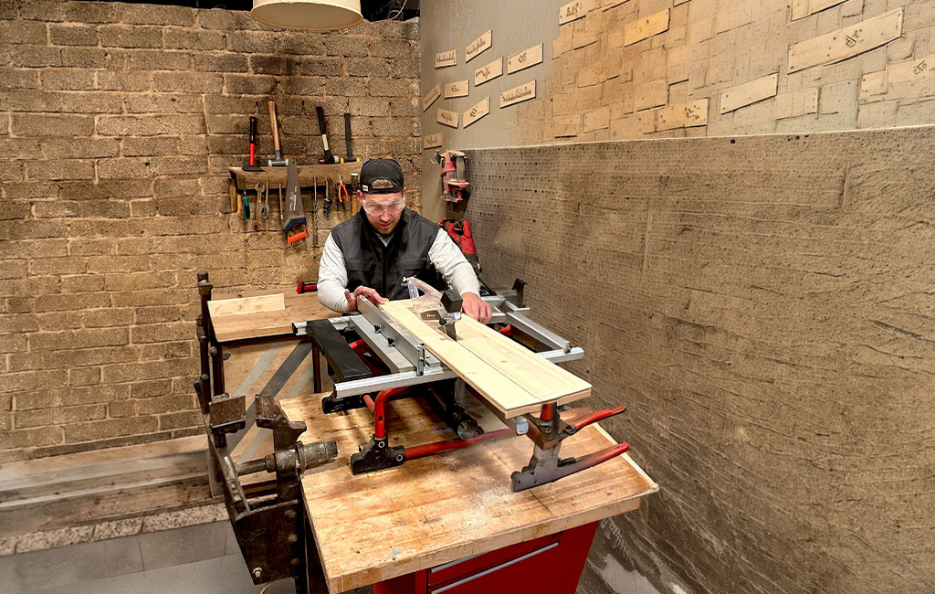 Person pushing a wooden board through a table saw in a workshop, cutting the wood with the visible circular saw blade on the machine.