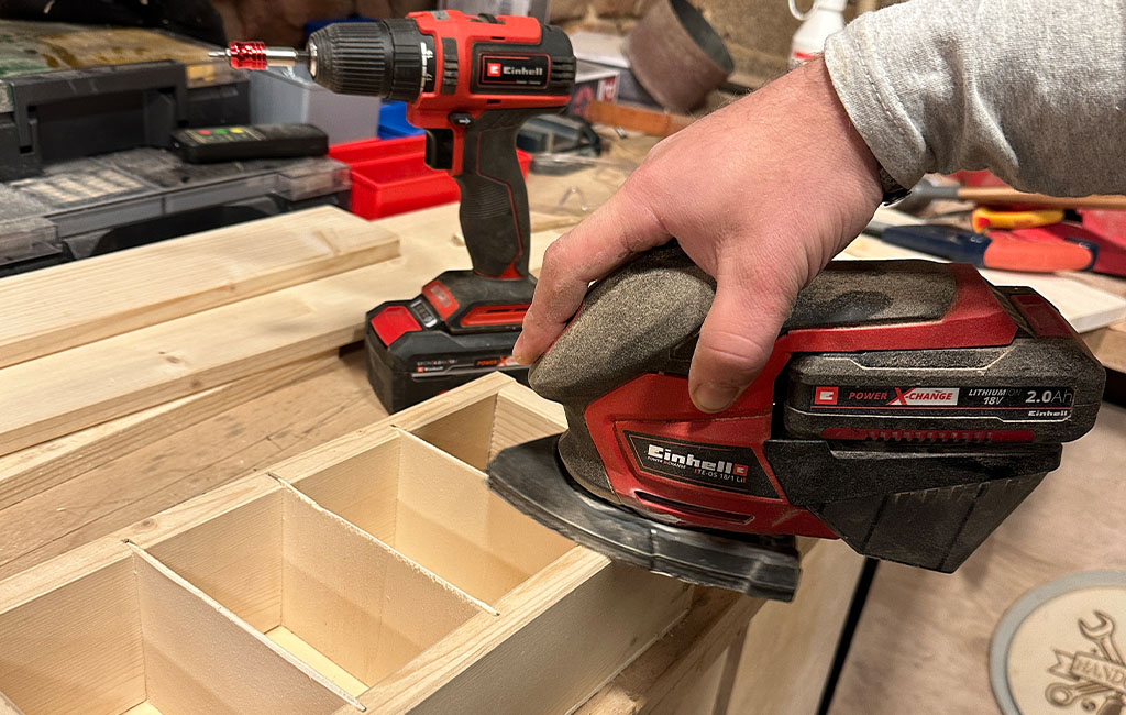 Person sanding the edge of a wooden box with a cordless orbital sander on a workbench, smoothing the surface during assembly.
