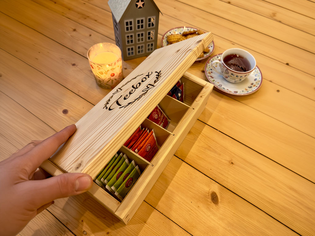 Handmade wooden tea box with engraved “tea box” lid and sorted tea bags inside, placed on a wooden table next to a cup of tea and candle.